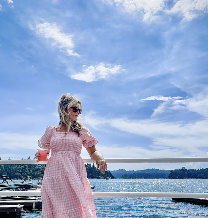 pink and white checkered maxi dress with smocked bodice and big puffy sleeves, maegan tintari, owner of dogwood dockside standing on the dock at the lake arrowhead marina in a pink dress and white crocs