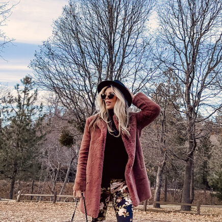 casual 70s boho look-floral bell bottom flares-faux fur coat-blonde woman standing on a tree-lined street with fall colors and leaves and blue cloudy skies