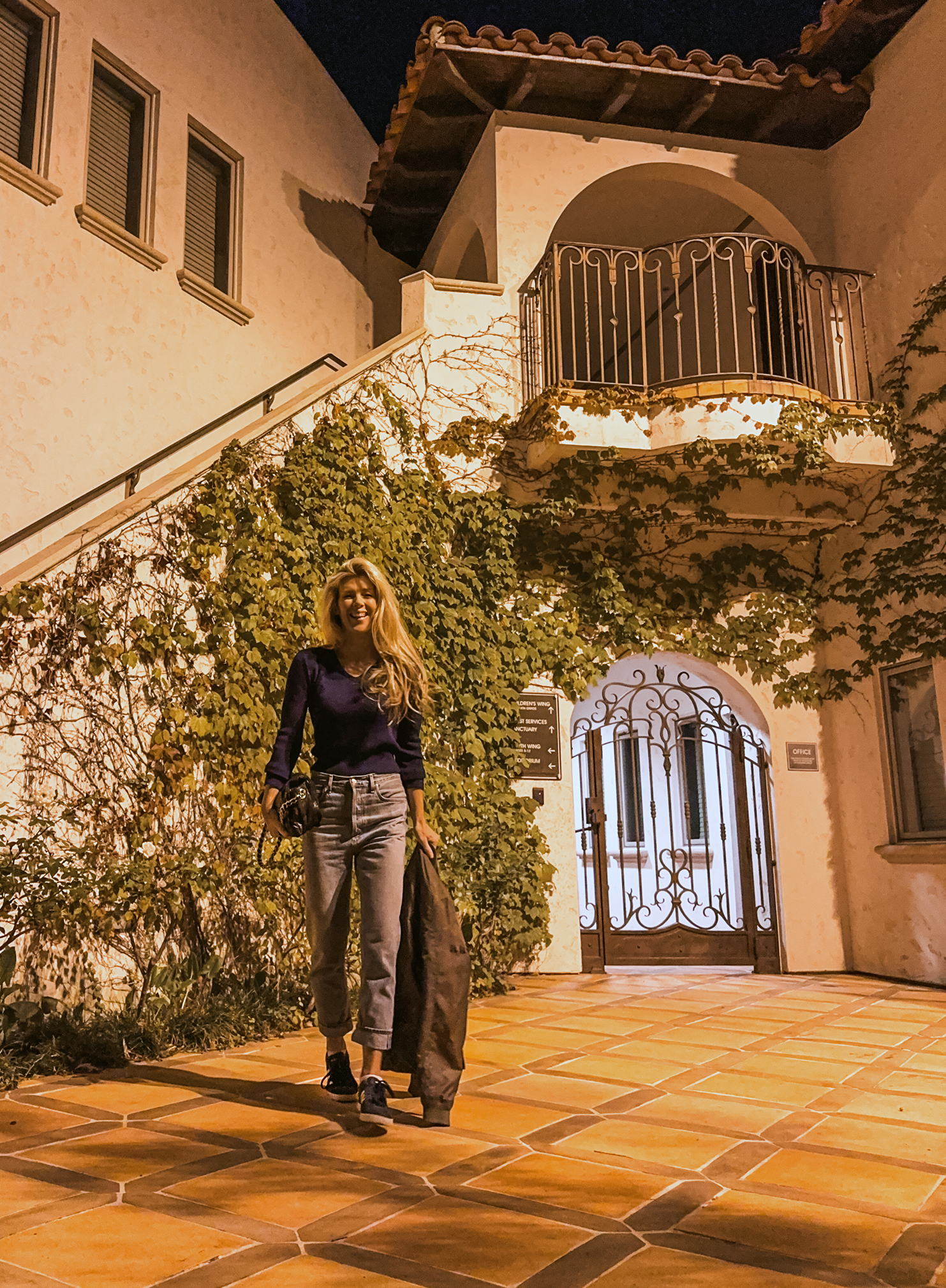 casual style light wash jeans and vintage short v-neck sweater with Adidas standing in front of a Spanish style house building at night