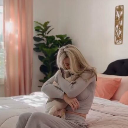 blonde woman hugging a pet french bulldog sitting on a pink and peach bed with pink curtains wearing grey matching set