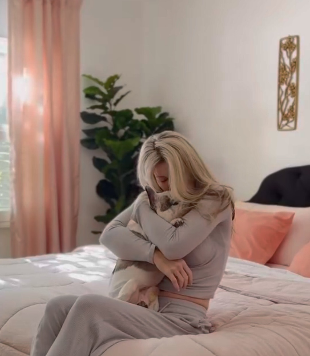 blonde woman hugging a pet french bulldog sitting on a pink and peach bed with pink curtains wearing grey matching set