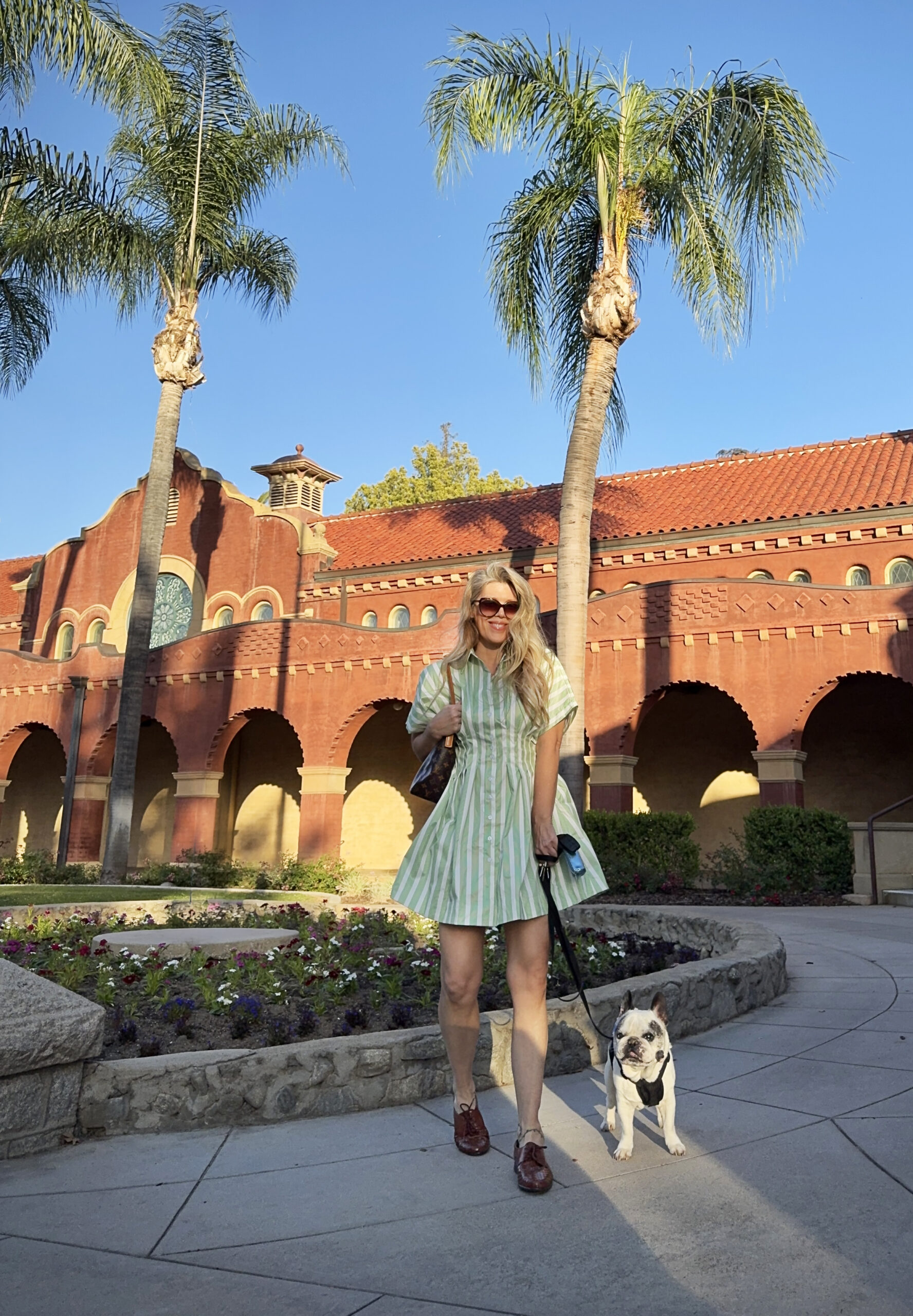 feminine style fashion blogger Maegan Amore walks her french bulldog, Trevor, in front of a Spanish style building blue sky and palm trees in the background wearing a cute green and white striped mini shirt dress with lace up oxfords and a louis vuitton purse